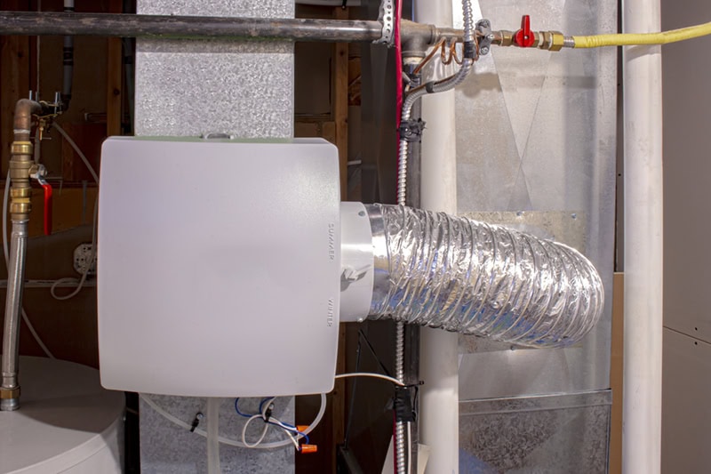 A white whole-house humidifier unit installed on sheet metal HVAC ductwork, connected via a flexible insulated foil duct and nearby copper piping.
