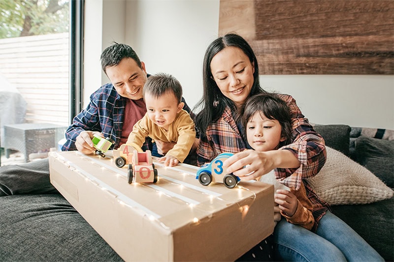 A family of four sitting together indoors on a sofa, smiling as the parents play with numbered toy cars on a cardboard box track with their two young children.
