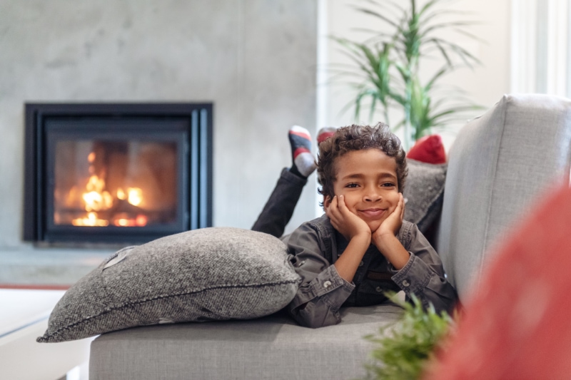 Gas Fireplaces - Staying Safe. Image is a photograph of a little boy laying on his belly on a grey couch and resting his chin in his palms in front of a gas fireplace.