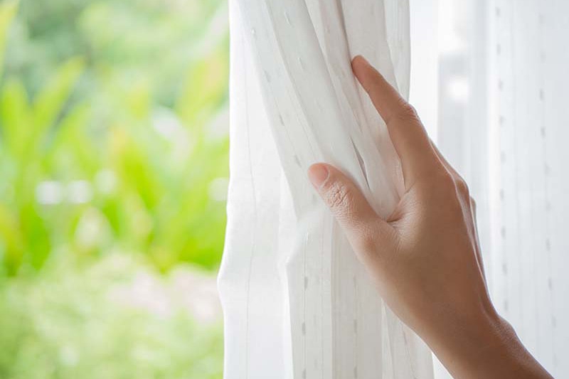 Dryer Vent Cleaning. Image is a photograph of a person's hand opening a window curtain with natural light and a garden background.